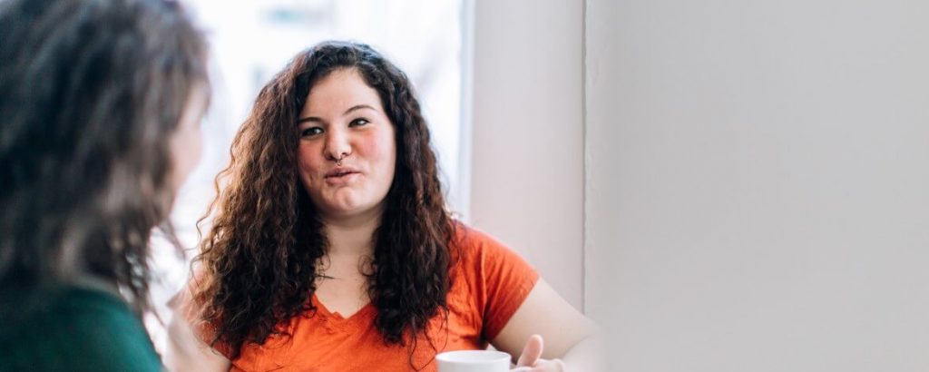 Image of a woman with curly brown hair facing another woman, talking over coffee