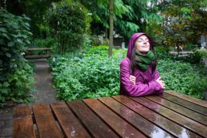 a young woman wearing a purple raincoat and green scarf, smiling peacefully at a picnic bench on a rainy day