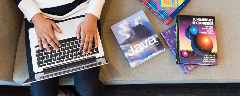 Image of a woman typing on a laptop while seated next to multiple textbooks on couch