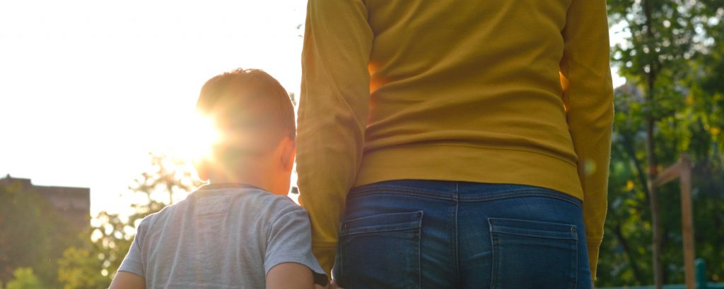 Image of a little boy holding his mother's hand, looking toward and playground on a sunny day