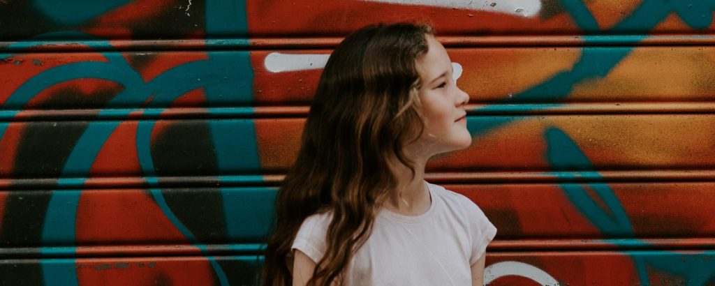 Image of a girl with long wavy hair, standing in front of a wall of graffiti